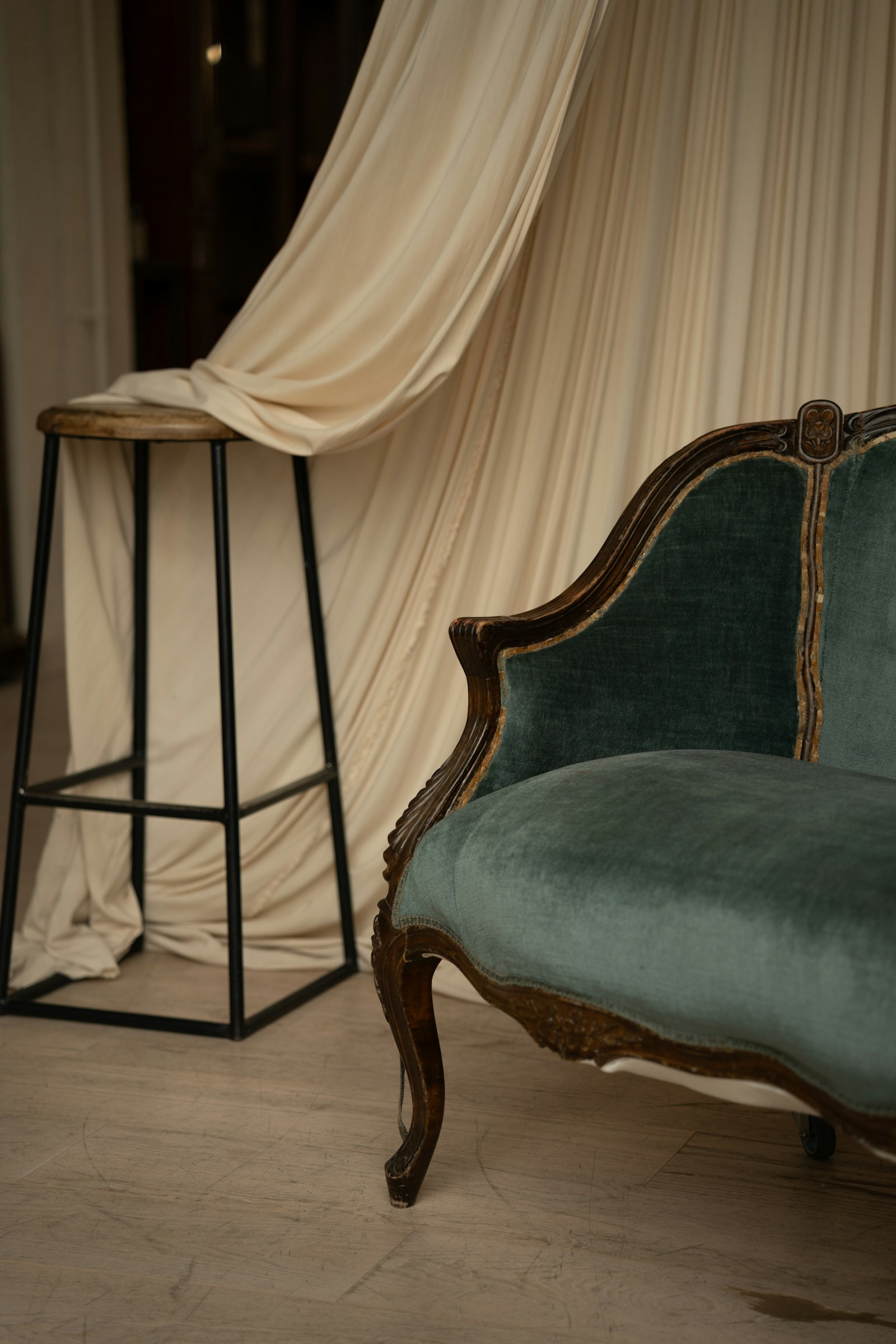 A carved-wood settee upholstered in muted teal velvet against a draped cream backdrop